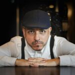 Close-up portrait of a man wearing a cap and white shirt, leaning on a table in a dimly lit room.